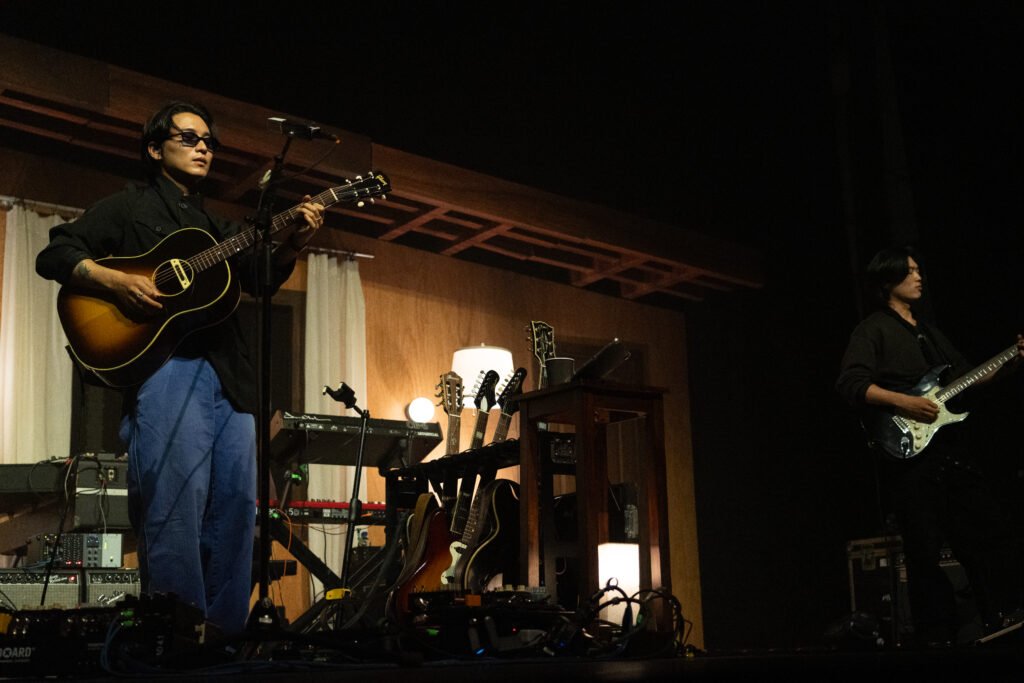 Two members of wave to earth perform with guitars inside a house-inspired stage set.