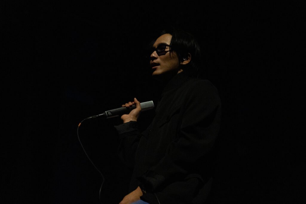 A wave to earth member holds a microphone and speaks to the audience under dim stage lighting.