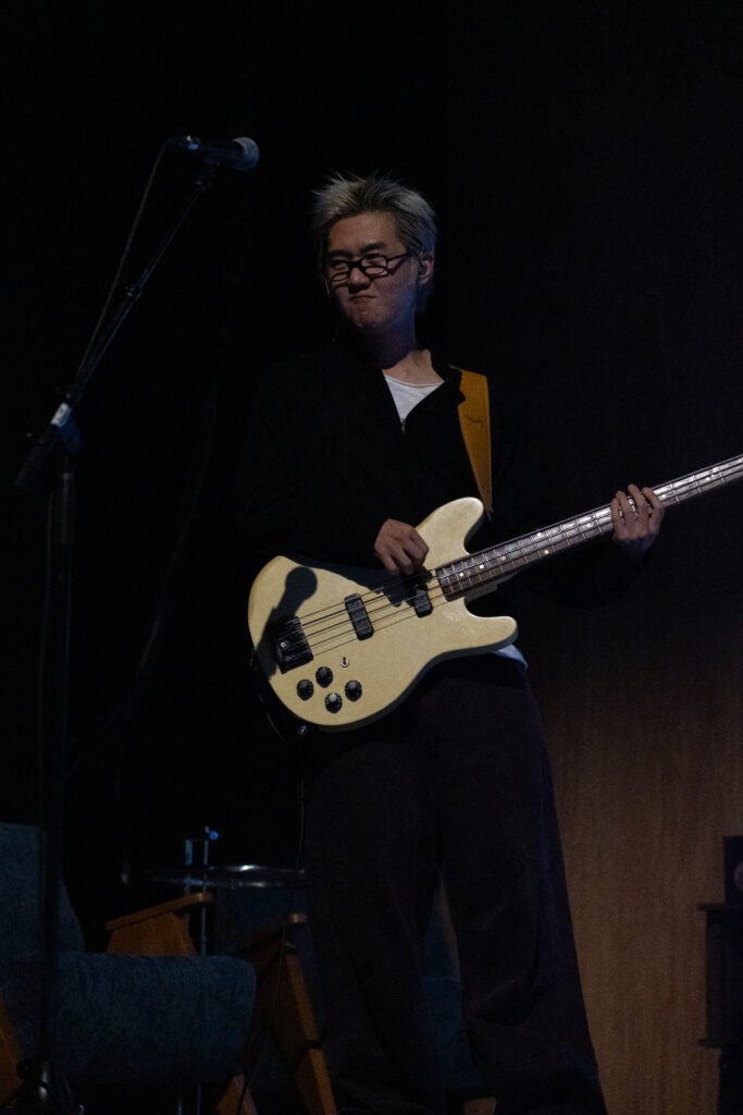 A bassist from wave to earth performs with a cream-colored electric bass under dim stage lighting.